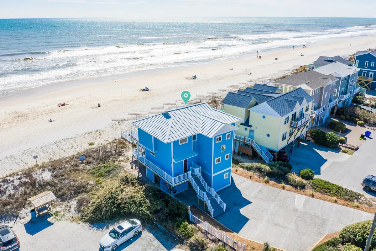 This exterior shot captures a beachfront property with ocean views, colorful houses, sand dunes, and parked cars visible in the scene. The vibrancy and proximity to the beach evoke the spirit of adventure and relaxation, further emphasizing arrival by car and easy access to the shore.