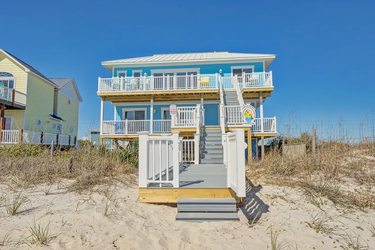 The relaxed and cheerful exterior of a blue beach house, featuring white railings, stairs leading up to a deck, and surrounding sand and grass. The setting showcases sunrise and sunset views from the deck and immediate access to outdoor activities like lounging and gathering with family.
