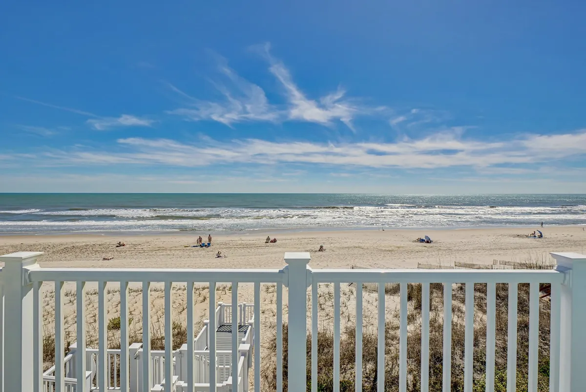 A beautiful view of the ocean waves and sandy beach, with people sunbathing and playing under a blue sky. A white railing in the foreground suggests proximity to a private deck or porch.