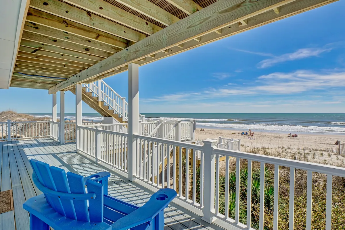 A blue Adirondack chair sits on a wooden deck with a clear view of the beach and gentle waves, framed by a sturdy railing and stairs leading directly to the sand. The scene radiates tranquility and offers a sense of seamless indoor-outdoor living.