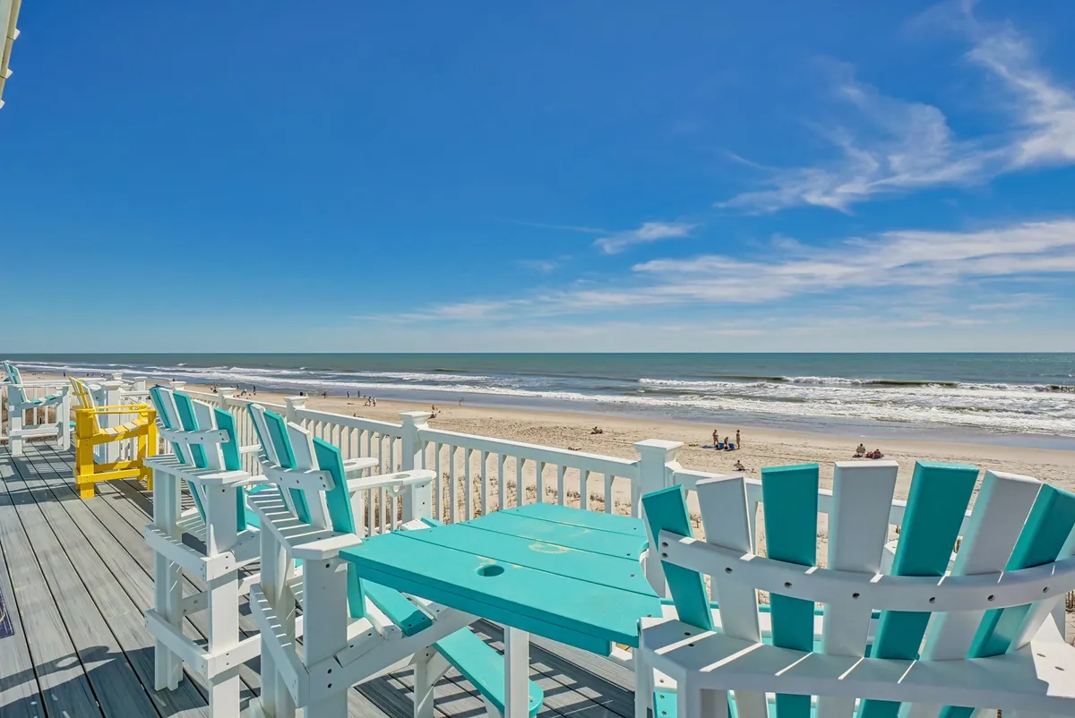 The image showcases a spacious deck lined with colorful Adirondack chairs overlooking sweeping views of North Topsail Beach and the sparkling ocean waves. The scene is set for relaxation and social gatherings, with the sun casting a inviting glow—perfect for sunset dinners, morning yoga, or simply soaking in the tranquility of the shore.