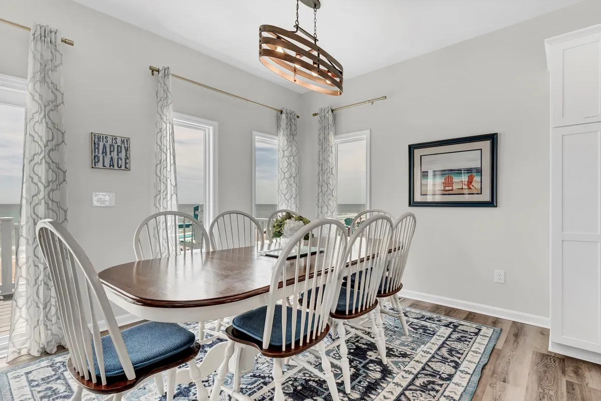 A stylish dining area with a large table set beneath a chandelier, surrounded by windows that frame panoramic ocean views. A decorative sign adds personality, and the table is clearly suited for hosting family dinners or festive gatherings, with a cozy, inviting atmosphere.