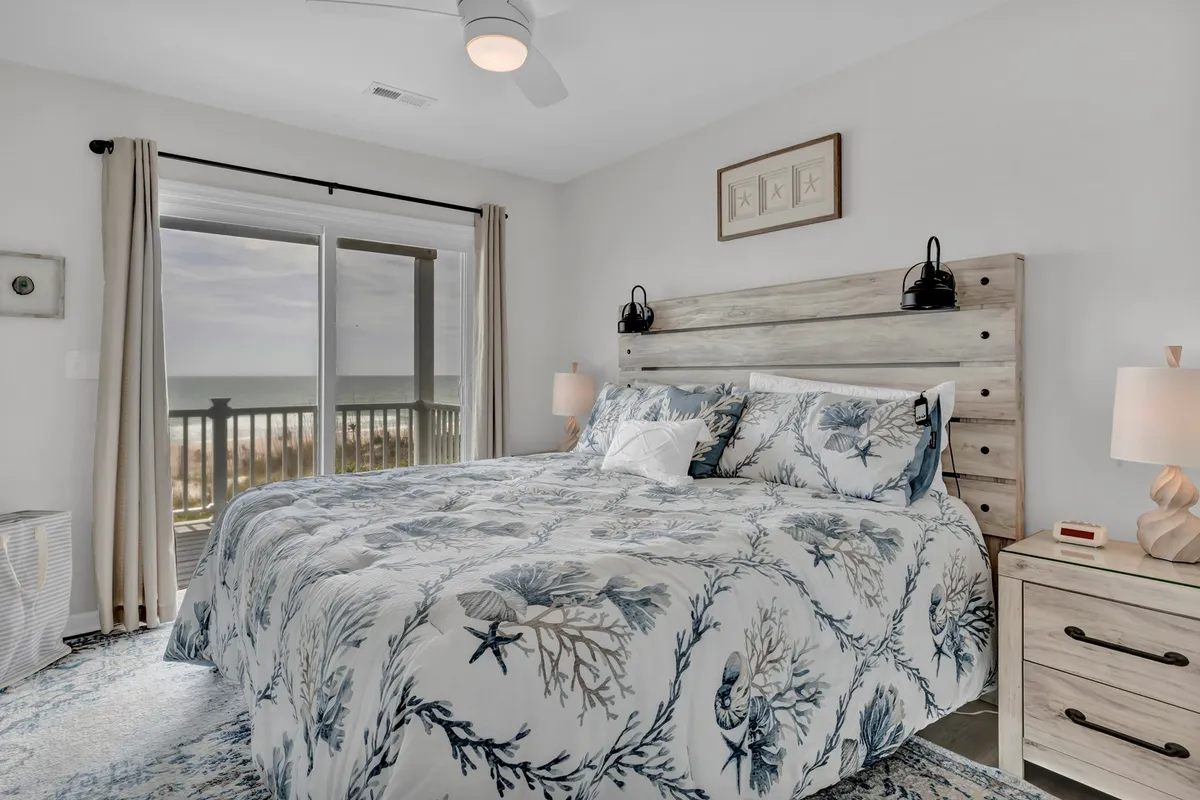 A serene bedroom featuring a large bed adorned with intricate bedding, flanked by a nightstand and lamp. Through the window, you can see an ocean view, enhancing the calming atmosphere. Soft lighting and subtle decor create a peaceful, spa-like ambiance.