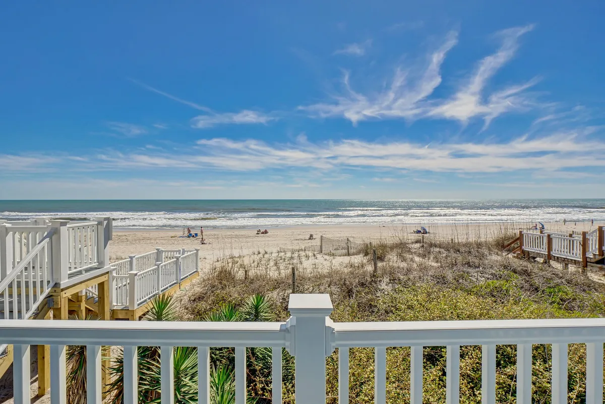 A tranquil beachfront view showing the vast ocean, sandy dunes, a wooden railing leading down to the beach, and a wide expanse of sky. The scene evokes classic summer activities such as sunbathing, swimming, and walking along the shore, with plenty of space for picnics and beach games.