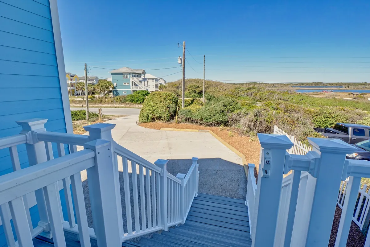 This image features the exterior of a blue house, a staircase leading up to the main entrance, surrounded by greenery and with a view of neighboring houses. The ample parking area is visible, suggesting convenience for guests arriving by car. The welcoming atmosphere hints at gathering spots and easy access to the beach and nearby amenities.