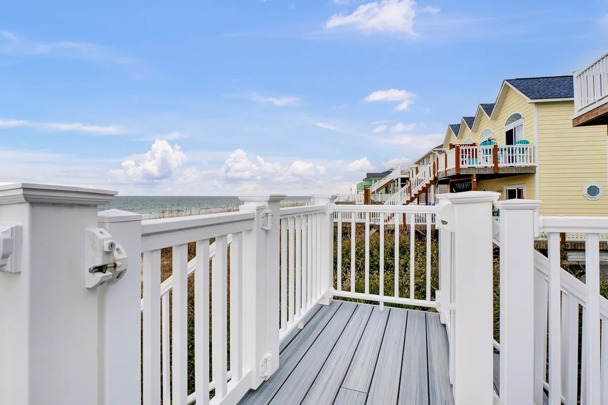 An exterior view from a deck with a railing, overlooking the ocean with neighboring houses in sight. The setting sun and the peaceful environment suggest opportunities for relaxation, sunset viewing, and hosting barbecues after a day of exploring the island.