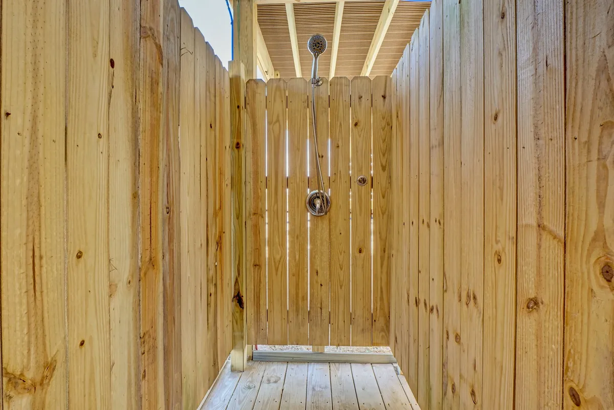 A wooden outdoor shower with a modern showerhead stands enclosed by a fence, bathed in natural light. The setup is inviting, perfect for rinsing off after sandy beach outings while enjoying the coastal breeze.
