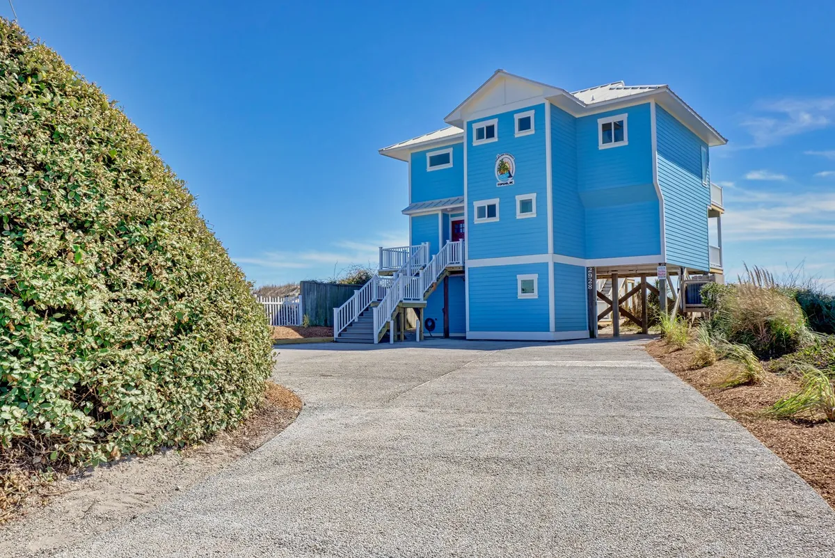 This image captures the cheerful blue house exterior with a prominent staircase leading up from the sandy landscaping. The scene conveys easy access from the house to the outdoors, suggesting a seamless transition for guests and their pets after a beach adventure.