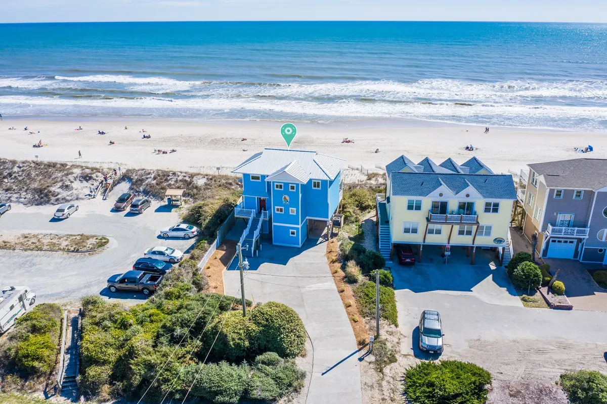 A striking blue beachfront house set against the ocean, with a sandy beach and gentle waves visible, surrounded by other coastal homes. The expansive beach area invites lounging, games, and water sports while the home’s elevated deck provides unobstructed sunrise and sunset views.