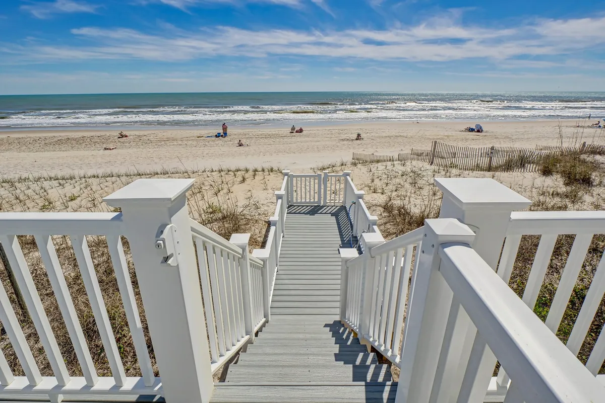 A wide exterior shot showing a staircase leading down to the sandy beach, with ocean waves rolling in and families relaxing on the sand. The deck overlooks both the shoreline and people gathered for leisure activities.