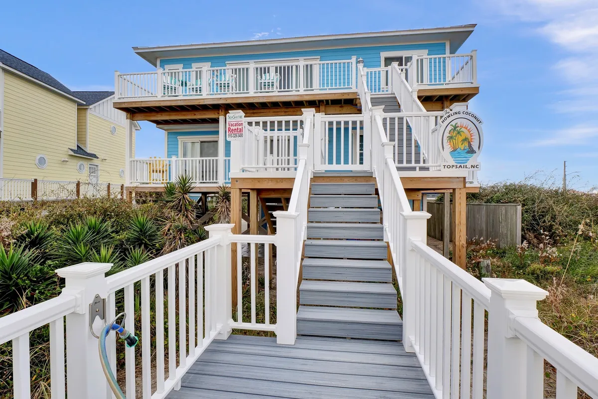 The blue house with a prominent wooden staircase and deck, complemented by a sturdy railing and open space that transitions directly to the beach. The scene captures outdoor relaxation, family gatherings, and the seamless connection to nature.