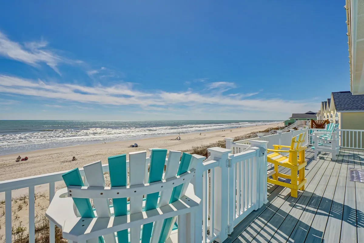 A beachfront deck adorned with colorful chairs invites relaxation under a vast blue sky, with unobstructed ocean views stretching out beyond the wooden railing. The seating area is positioned for enjoying sunrise coffees, sunset cocktails, or simply soaking up the tranquil seaside atmosphere.