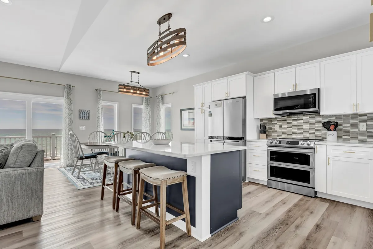 A modern kitchen space with a large island bar, stainless steel Samsung Smart appliances, ample windows allowing natural light and water views, and decorative lighting. Bar stools along the island invite casual breakfasts or group meal prep, and the overall design feels bright, functional, and upscale.