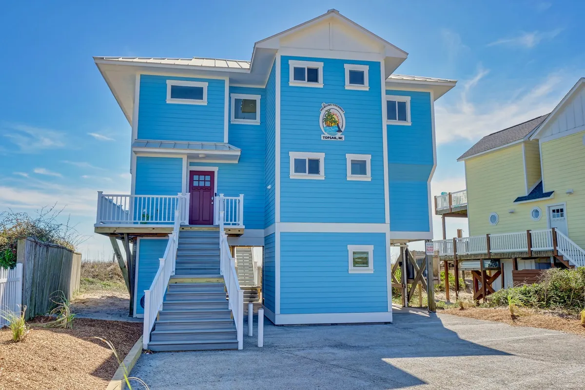 The cheerful exterior of a bright blue house with a staircase leading to the entrance and a decorative sign. Family members are seen gathering on the porch, with easy access to beach outings and group relaxation.