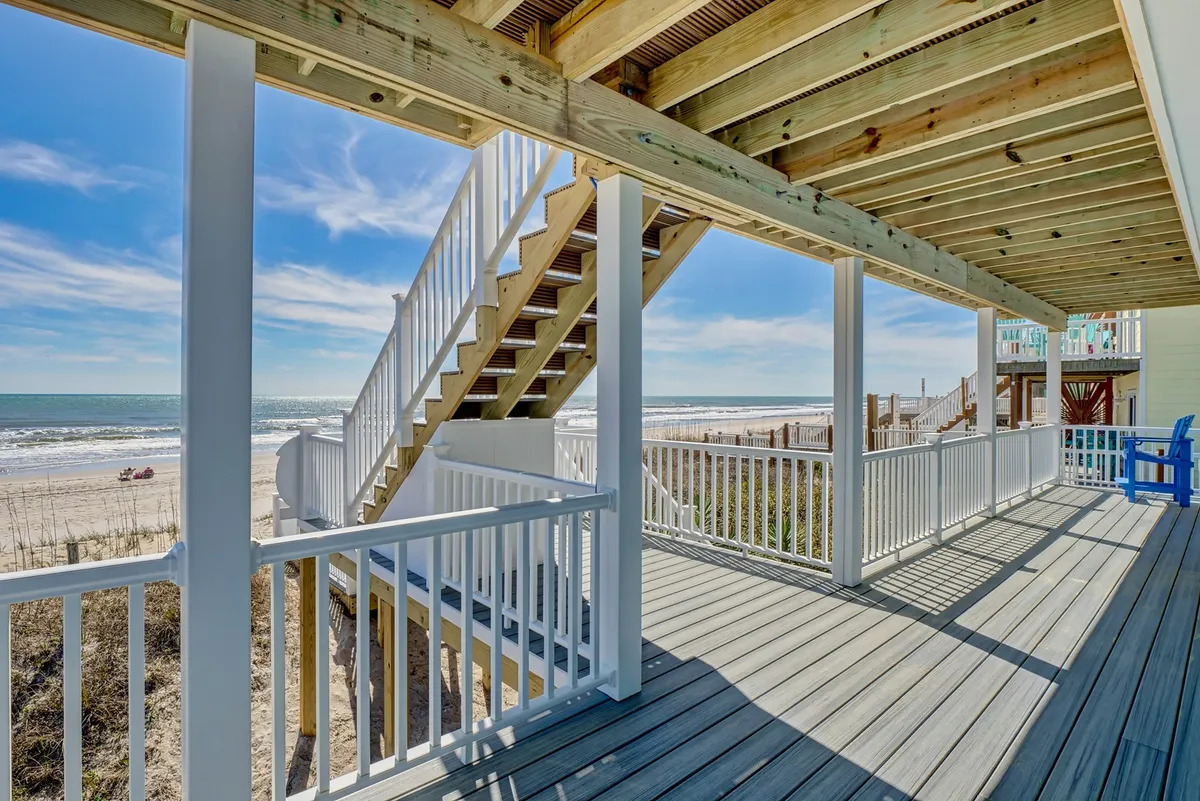 A relaxed deck scene with wooden railings, comfortable outdoor furniture, and a sweeping view of the beach and ocean just beyond. The atmosphere hints at morning coffees, sunset wine, and family gatherings overlooking the water.