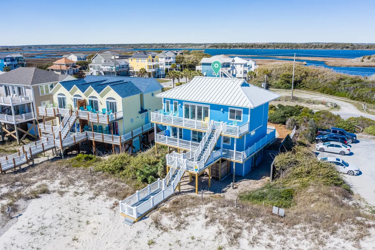 An exterior shot of the blue house with staircase, surrounding beach, parking area, and nearby water. Multiple cars can be seen, giving a clear sense of the ample parking and proximity to the beach, key for guests arriving with pets or multiple vehicles.